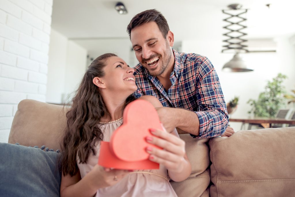 Jeune femme souriante ouvrant la boîte de chocolats que lui offre son amoureux, sur le canapé du salon. 